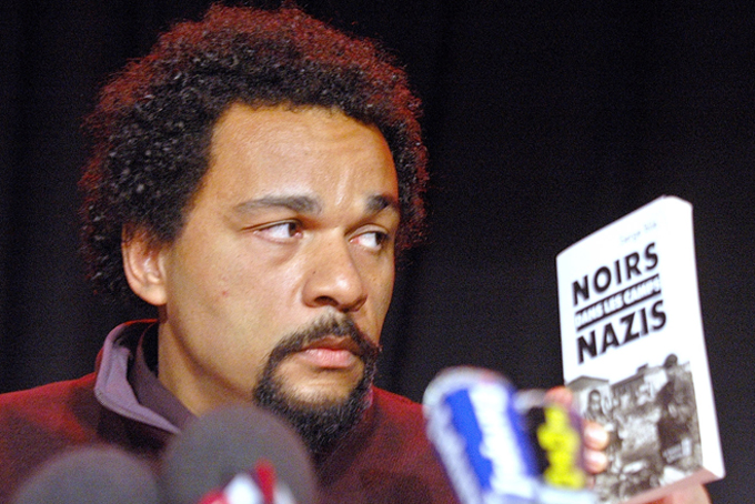In this Feb. 19, 2005 file photo, French comic Dieudonne M'Bala M'Bala looks at a book "Blacks in the Nazi camps" by Serge Bile during a statement at La Main d'Or theater, in Paris. (AP Photo/Jacques Brinon, File)