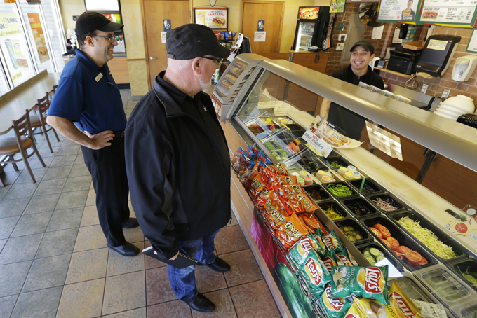 David Jones, center, talks with his employee Roberto Castelan, right, and regional manager Chris Rodrigues, left, during a visit to one of the two Subway sandwich franchises Jones owns, Tuesday, March 3, 2015, in Seattle. Jones, who has 18 employees at his two stores, says he will need to raise prices 4 percent to cover the first pay increase mandated by Seattle's minimum wage law. (AP Photo/Ted S. Warren)