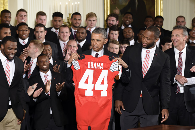 President Barack Obama holds up a Ohio State football jersey that he was presented with from Ohio State football players Curtis Grant and Doran Grant, as head coach Urban Meyer watches at right, as he welcomed the NCAA College Football Playoff National Champion Ohio State Buckeyes, Monday, April 20, 2015, during a ceremony in the East Room of the White House in Washington. (AP Photo/Susan Walsh)