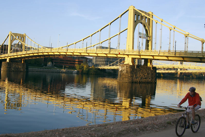 The setting sun adds a warm glow to the yellow of the Roberto Clemente Bridge as a cyclist pedals by on the trail on the southern shore of the Allegheny River on Monday, Oct. 28, 2013, in Pittsburgh. (AP Photo/Keith Srakocic)
