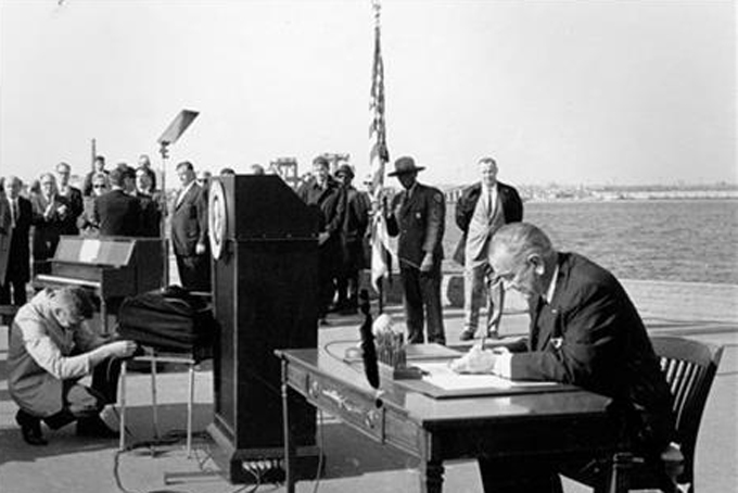 FILE - In this Oct. 3, 1965, file photo President Lyndon B. Johnson sits at his desk on Liberty Island in New York Harbor as he signs a new immigration bill. It was presented as a symbolic move - President Lyndon Johnson on Liberty Island, signing an immigration bill that gave people from every country in the world an equal chance to come to America. Fifty years later, there’s little that hasn’t been changed as a result of the Hart-Celler Act that Johnson signed on Oct. 3, 1965. A country that was almost entirely native-born in 1965 has a significant foreign-born population; demographic diversity has spread to every region, expanding a black-and-white racial paradigm into a multi-colored one. Americans have gleefully adopted musical genres and foods that have immigrant origins, while remaining conflicted and uneasy politically over who’s here, legally and not. (AP Photo, File)