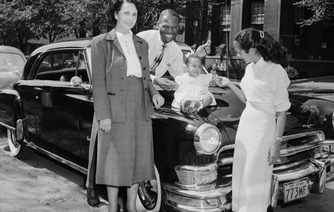 Charles “Teenie” Harris; Two women and man standing around baby girl seated on hood of dark Chrysler with Pennsylvania license plate number “773MF,” on Centre Avenue with YMCA in background, Hill District, c. 1951; Heinz Family Fund, Carnegie Museum of Art
