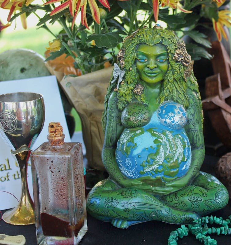 A green, ceramic, seated female figurine sits beneath a tree, with a metal wine glass and a bottle containing a red liquid placed beside her.
