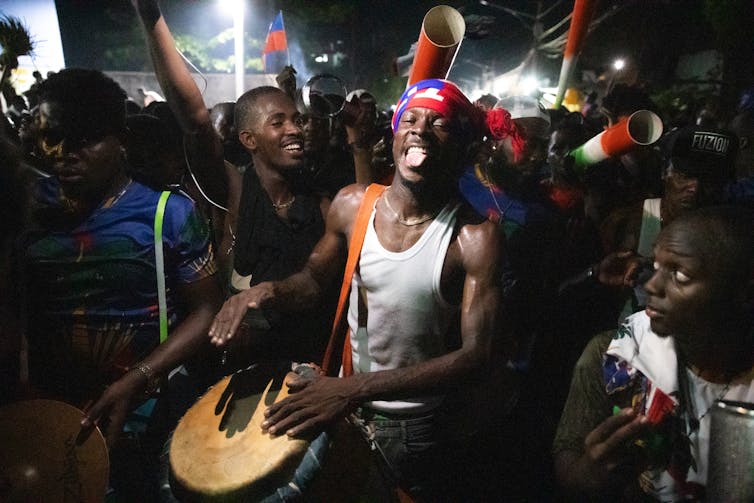 A man beats a drum surrounded by pther people