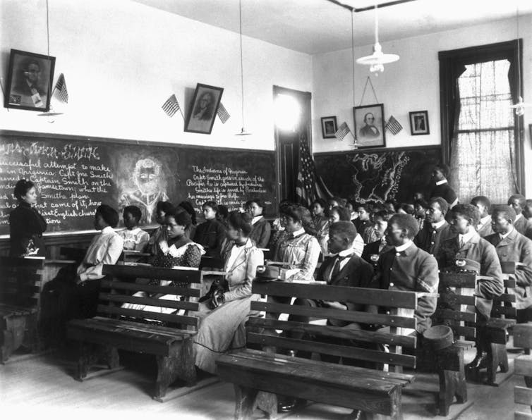 A black and white photo shows several rows of young Black people dressed formally sitting in rows of benches with a chalkboard nearby them.
