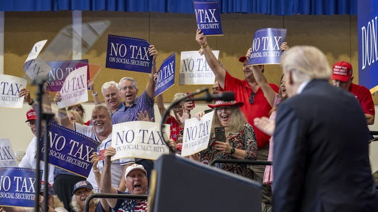 Older people waive signs that say 'No Tax on Social Security,' while Donald Trump stands at a podium.