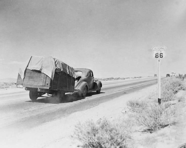Black-and-white photo of an old car hauling a trailer of belongings along a dusty stretch of highway.