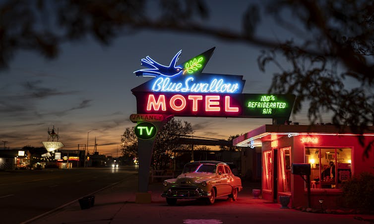 A roadside motel at dusk with a vintage car parked in the lot and a large blue and pink neon sign reading 'Blue Swallow Motel.'
