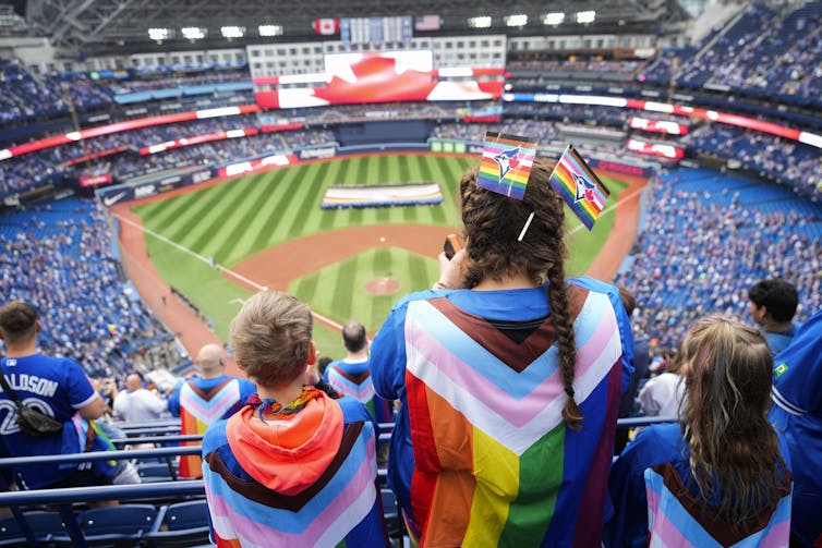 Fans wearing pride themed flags draped over their backs stand while facing the field, where a large pride flag is held up by volunteers
