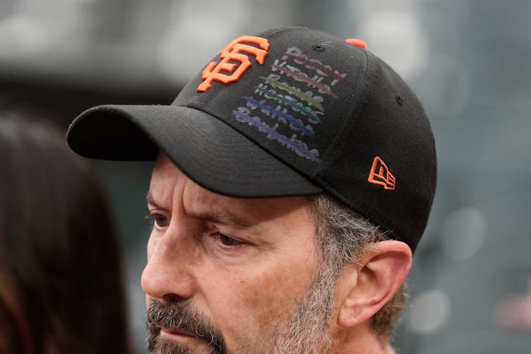 A man wearing a San Francisco Giants baseball hat with names written out in rainbow and crossed out